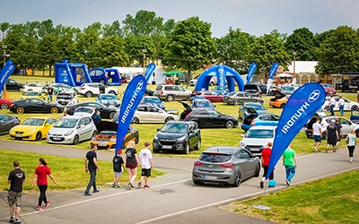 Hyundai Treffen Fotos | Motorsport Events Deutschland | Michael Schalansky Zahlreiche Hyundai-Modelle verschiedener Baureihen sind auf einem sonnigen, grasbewachsenen Showplatz aufgereiht, umgeben von Besuchern und Werbebannern. Das Bild wurde von Michael Schalansky aufgenommen.